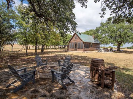 A small wooden cabin with a green roof, surrounded by trees, features an outdoor seating area with Adirondack chairs and a fire pit.