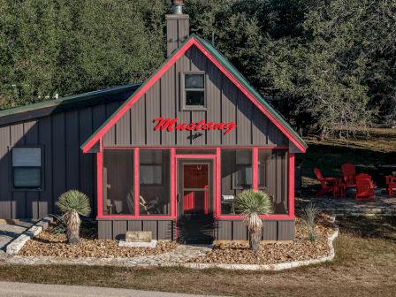 A small outdoor cabin with red trim labeled &ldquo;Mustang,&rdquo; potted plants, and red patio chairs by a stone path under trees.