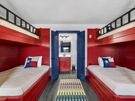 Two red-and-blue bunk beds with Texas-style flag pillows flank an open blue door into a small bathroom.