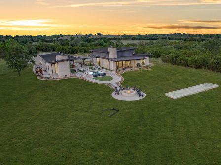 Aerial view of a modern luxury home with outdoor pool, patio firepit, and manicured lawn at sunset.