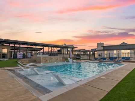 An outdoor swimming pool with lounge chairs and a landscaped courtyard at sunset, in front of apartment-style buildings.