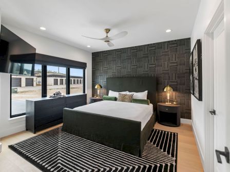 Modern bedroom with a king bed, dark textured accent wall, ceiling fan, bedside lamps, striped rug, and large windows.