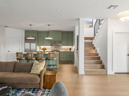 A bright open-plan living room leads to a green cabinetry kitchen and an indoor staircase in a modern home.