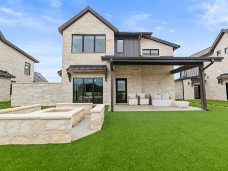 A modern stone house with large windows, green lawn, and an outdoor patio seating area under a metal pergola.