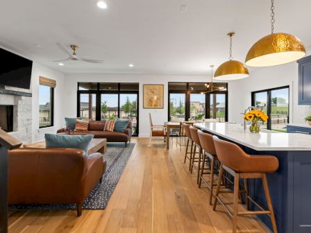 A modern open-plan living room and kitchen with leather sofas, bar stools, pendant gold lights, and large windows.