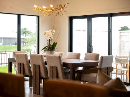 A modern dining room with a wooden table, beige chairs, chandelier, and white orchids by large windows, in soft light.
