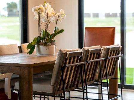 Sunlit modern dining area with wooden table and chairs, white orchid centerpiece, and hanging globe lights.