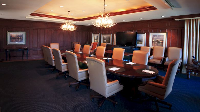 A conference room with a long table, leather chairs, wall art, two chandeliers, and a large screen on the wall; set up for a meeting with notepads.