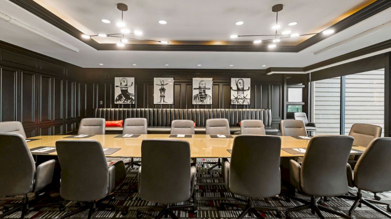 A modern conference room with long wooden meeting tables and rows of chairs, lit by recessed ceiling lights.