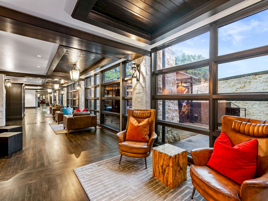 A modern hotel hallway lounge with leather armchairs, bright pillows, wood floors, and large windows overlooking a stone exterior.
