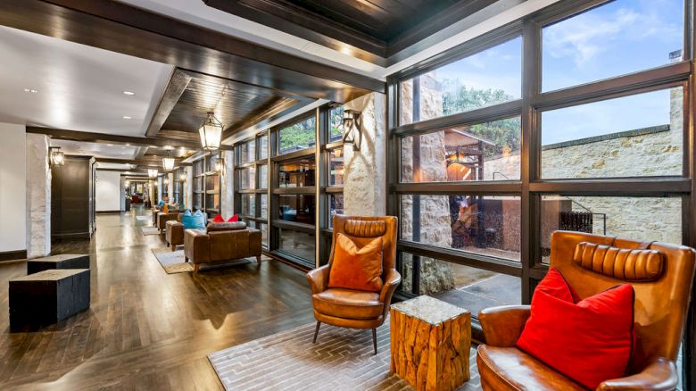 A modern hotel hallway lounge with leather armchairs, bright pillows, wood floors, and large windows overlooking a stone exterior.