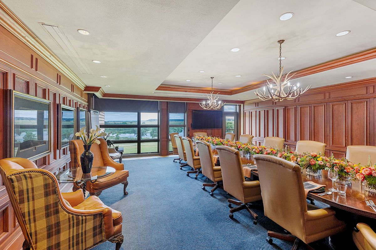 This image shows an elegant conference room with a long table, cushioned chairs, floral centerpieces, and large windows offering scenic views.