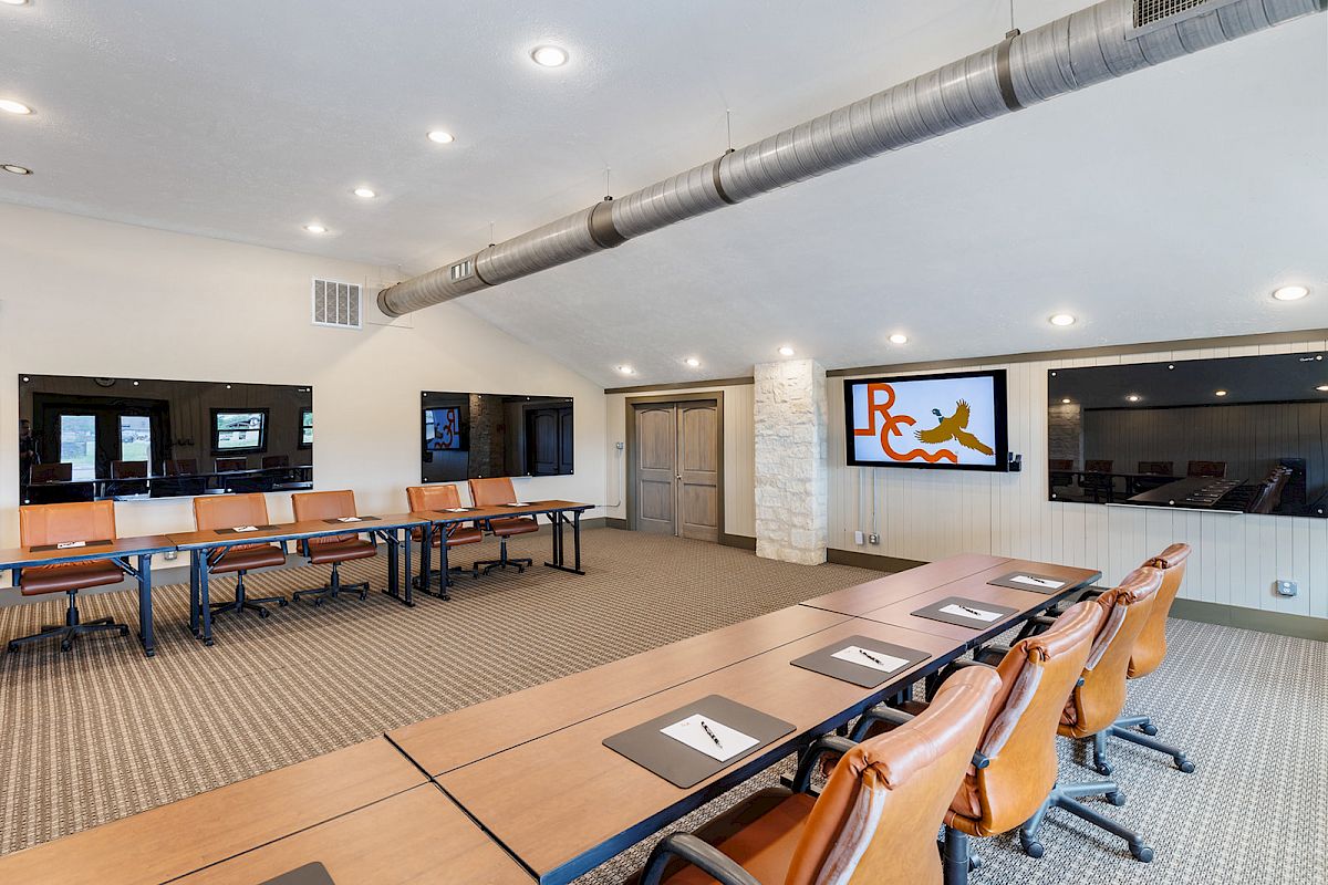 A modern conference room with long tables, chairs, and TV screens on the walls, featuring a logo on one screen.