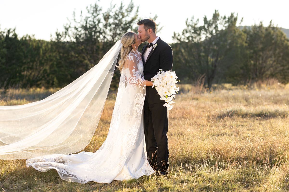A couple in wedding attire kisses in a meadow, with the bride in a long veil and gown holding a bouquet of white flowers.