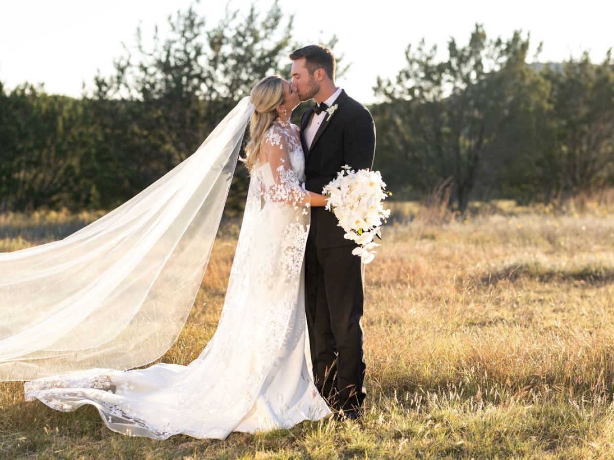 A couple in wedding attire kisses in a meadow, with the bride in a long veil and gown holding a bouquet of white flowers.