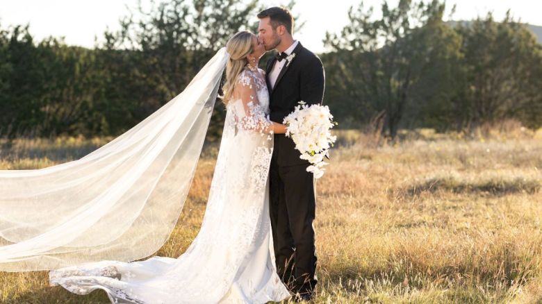 A couple in wedding attire kisses in a meadow, with the bride in a long veil and gown holding a bouquet of white flowers.