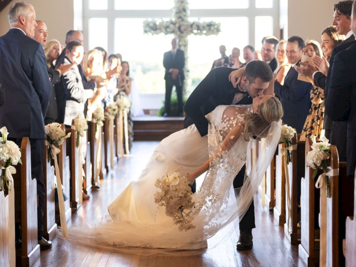 A couple kisses in a wedding ceremony while guests watch, captured in a church aisle adorned with flowers, in front of a windowed backdrop.