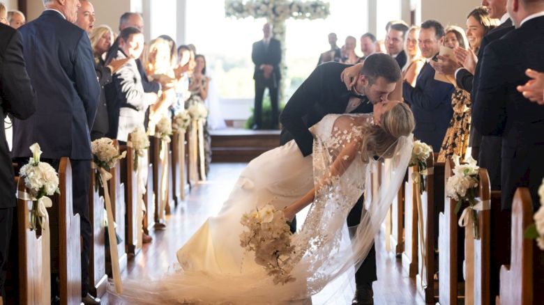 A couple kisses in a wedding ceremony while guests watch, captured in a church aisle adorned with flowers, in front of a windowed backdrop.