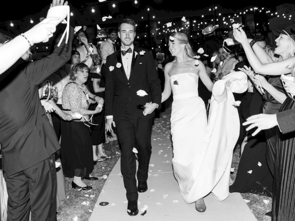 A newlywed couple walks down an aisle, surrounded by celebrating guests throwing petals at night, under string lights.