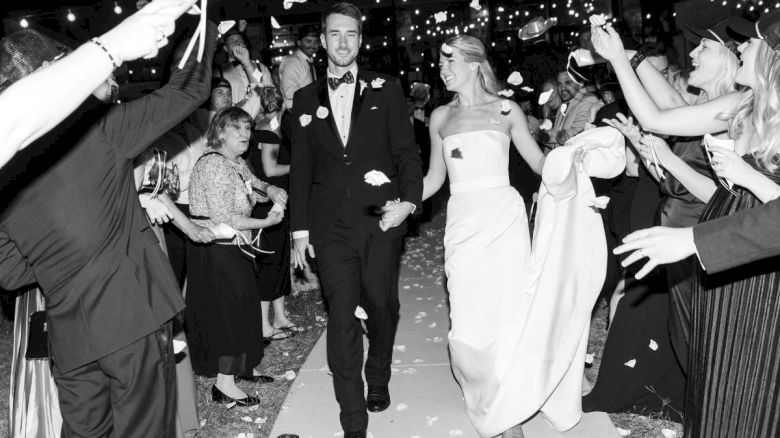 A newlywed couple walks down an aisle, surrounded by celebrating guests throwing petals at night, under string lights.