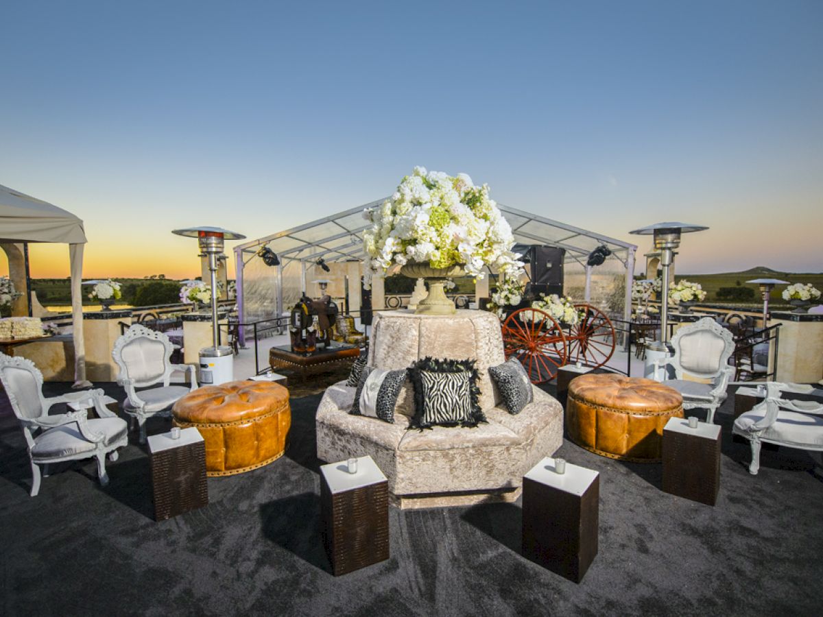 An outdoor event setup with elegant seating, plush furniture, decorative flowers, and tents under a sunset sky.