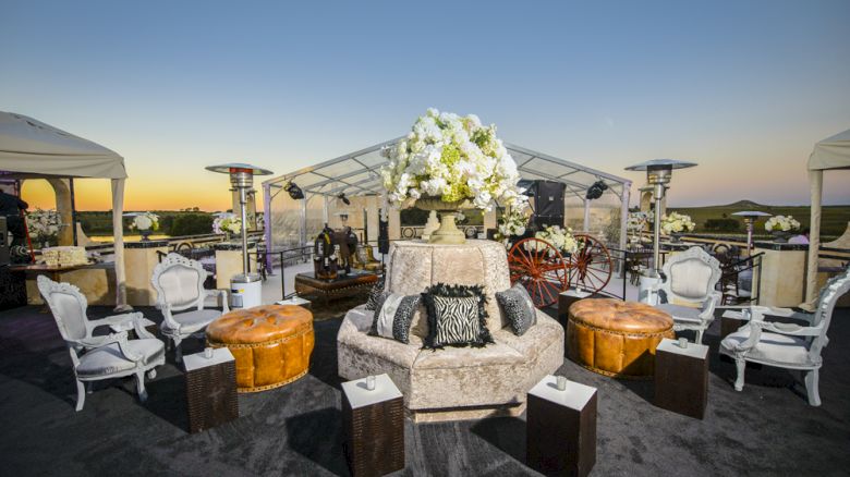 An outdoor event setup with elegant seating, plush furniture, decorative flowers, and tents under a sunset sky.