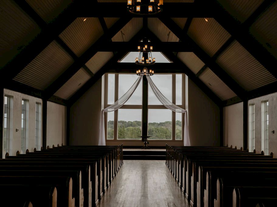 A quiet church interior with dark pews, tall arched windows, and hanging chandeliers framing the altar.