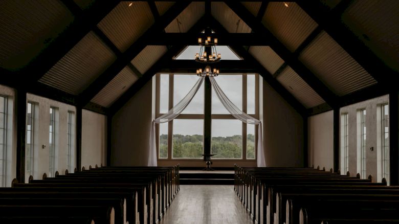 A quiet church interior with dark pews, tall arched windows, and hanging chandeliers framing the altar.