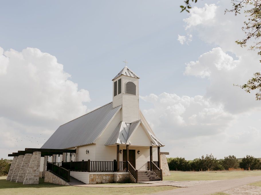 A white country church with a small bell tower and steps, standing outdoors under a cloudy sky.