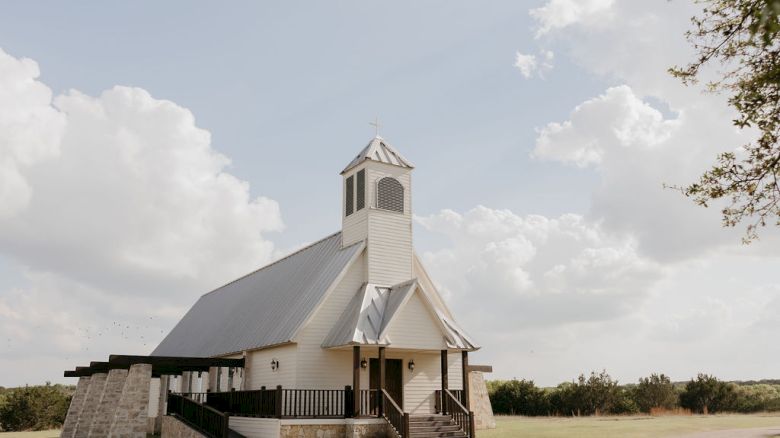 A white country church with a small bell tower and steps, standing outdoors under a cloudy sky.