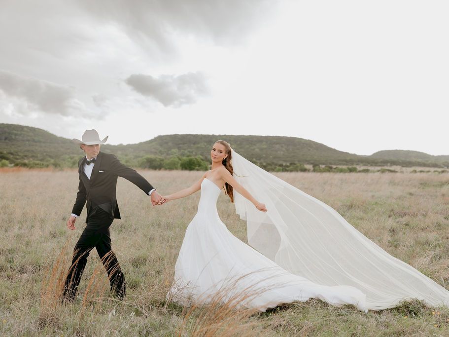 A bride in a white dress and veil walks hand-in-hand with a cowboy-hat groom across a grassy field.