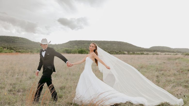 A bride in a white dress and veil walks hand-in-hand with a cowboy-hat groom across a grassy field.