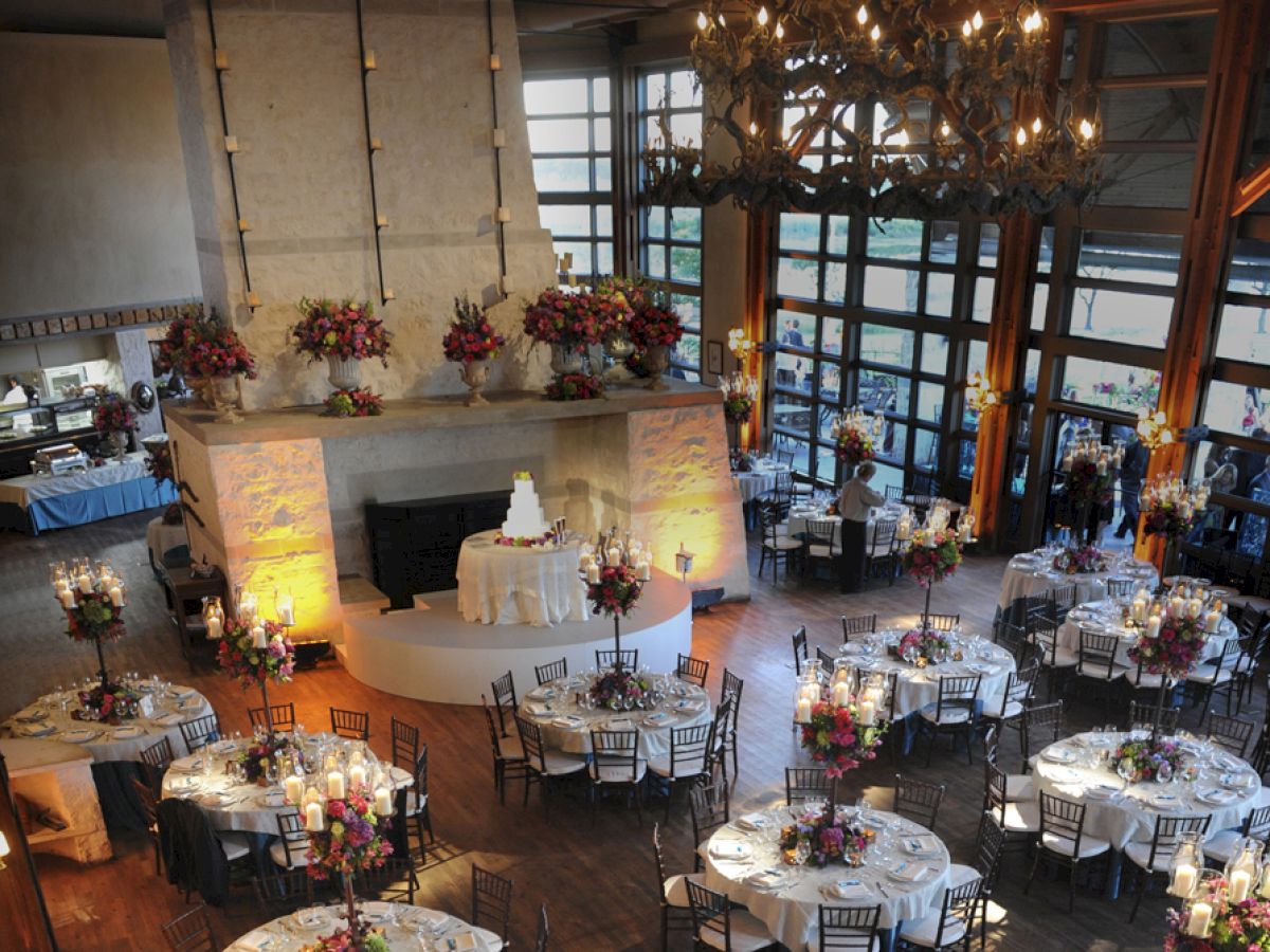 A decorated event space with round tables set for a reception, featuring floral arrangements and a central white tiered cake on display.