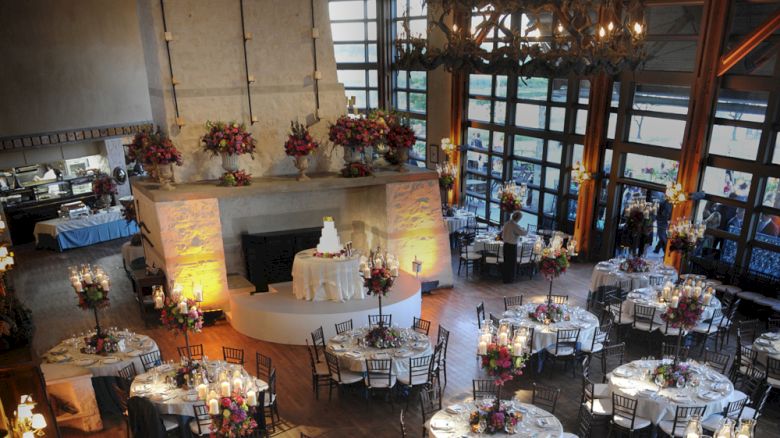 A decorated event space with round tables set for a reception, featuring floral arrangements and a central white tiered cake on display.