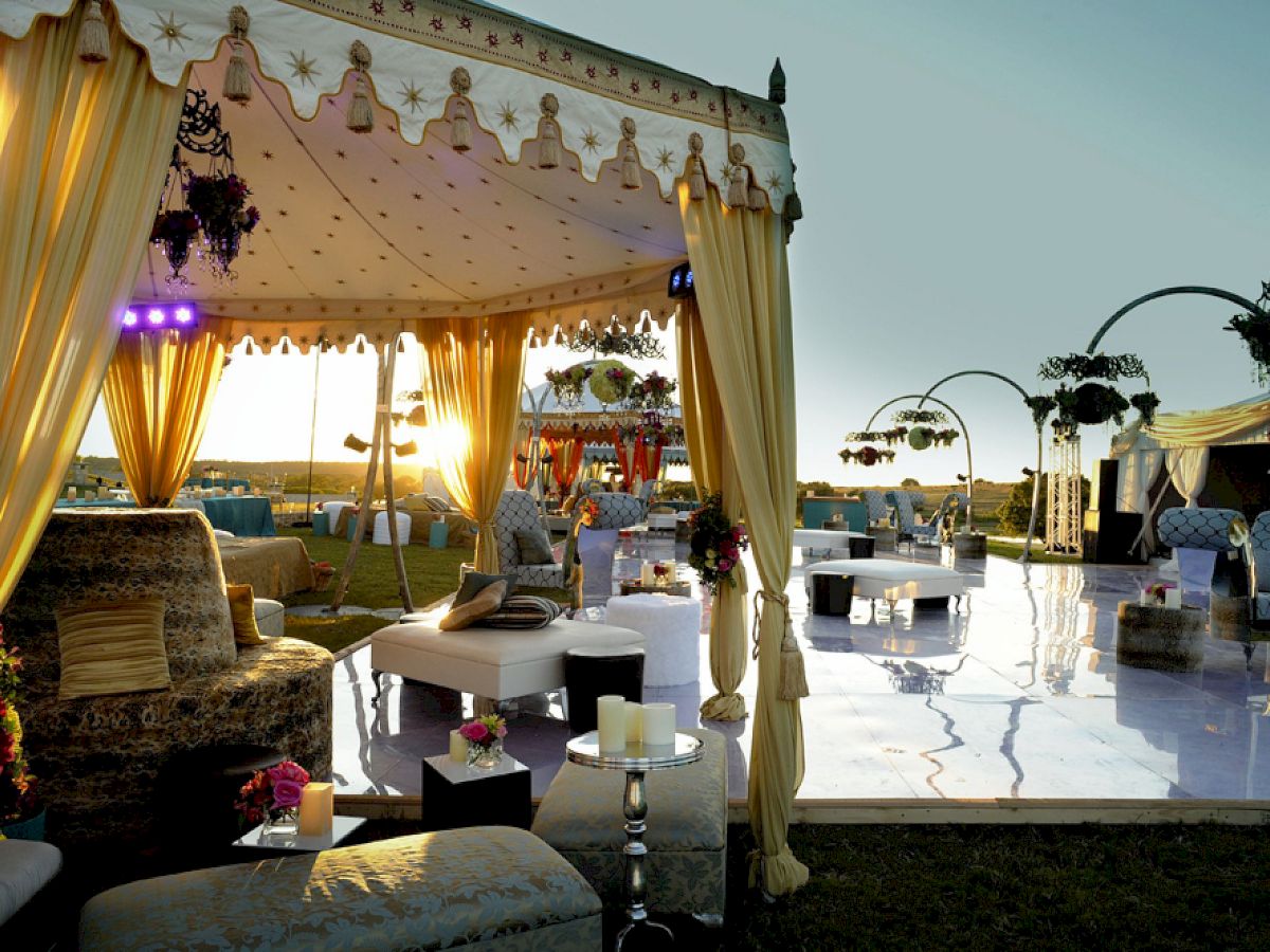 The image shows an elegant outdoor event setup with draped tents, luxurious seating, decorated tables, and lighting fixtures during sunset.