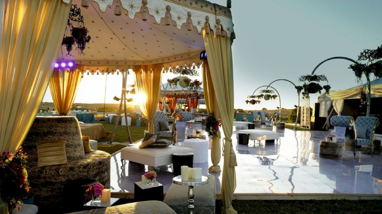 The image shows an elegant outdoor event setup with draped tents, luxurious seating, decorated tables, and lighting fixtures during sunset.