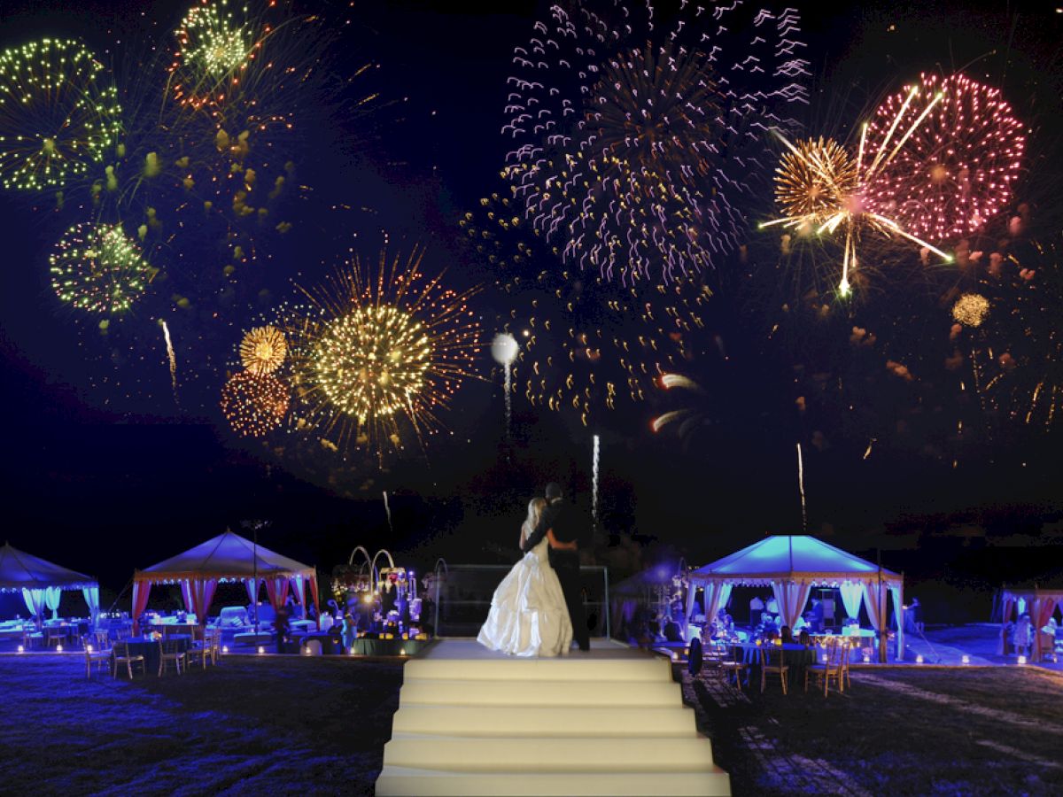 A bride in a white dress stands on steps, watching colorful fireworks in the night sky, with lit tents and guests in the background.