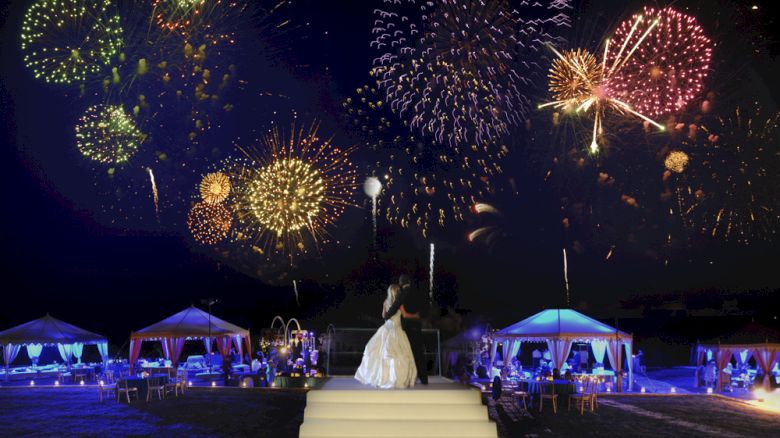 A bride in a white dress stands on steps, watching colorful fireworks in the night sky, with lit tents and guests in the background.