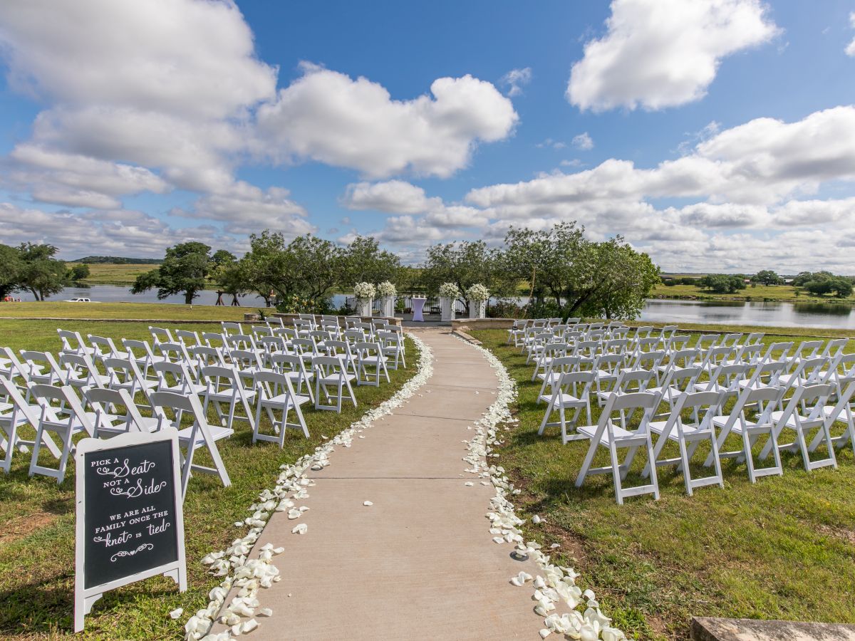 An outdoor wedding ceremony setup with white chairs down a walkway, flowers, and a lake under cloudy blue skies.