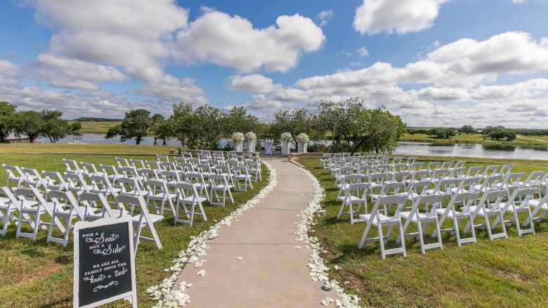 An outdoor wedding ceremony setup with white chairs down a walkway, flowers, and a lake under cloudy blue skies.