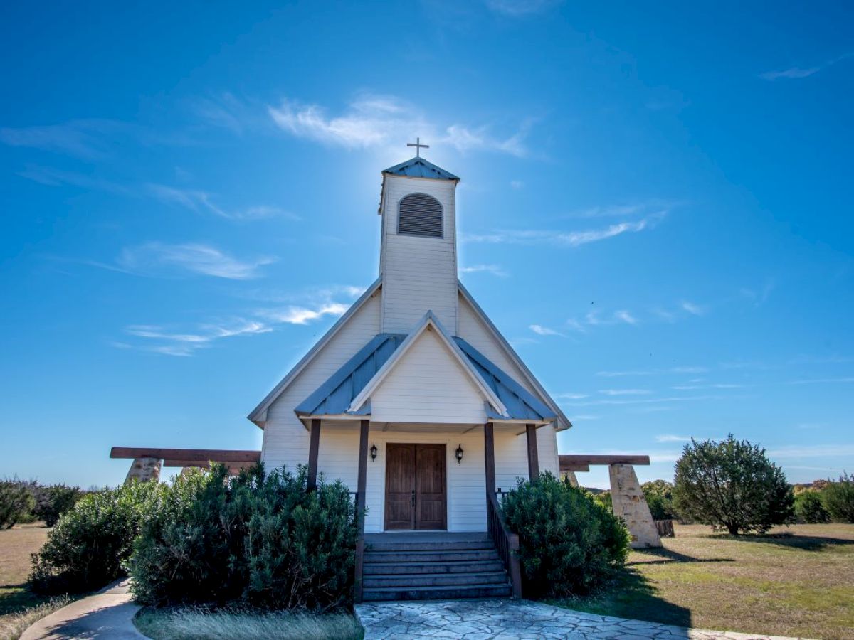 A small white church with a bell tower under a clear blue sky, surrounded by greenery and open fields, stands peacefully in the sunlight.