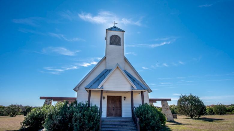 A small white church with a bell tower under a clear blue sky, surrounded by greenery and open fields, stands peacefully in the sunlight.