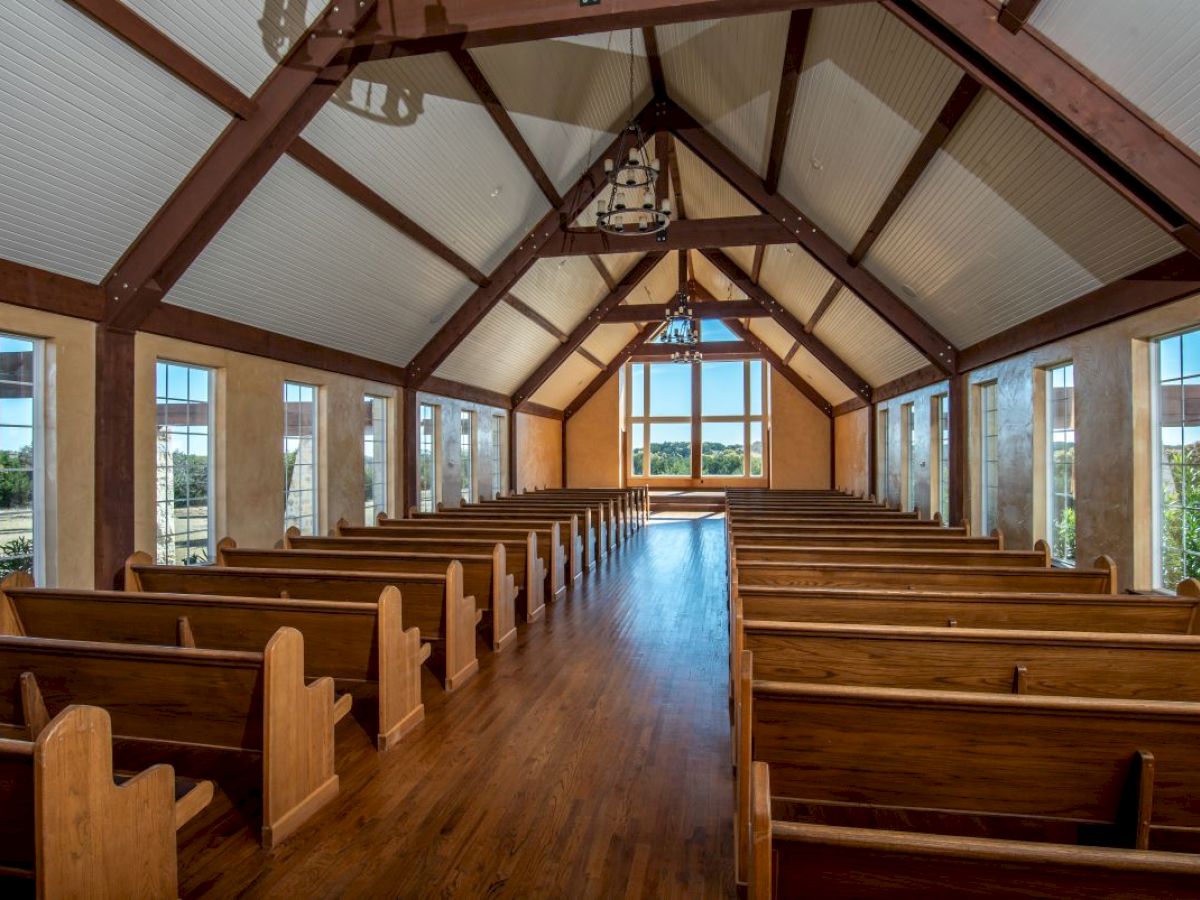 The image shows a wooden chapel interior with rows of pews, large windows, and a high ceiling, leading to a bright view outside.