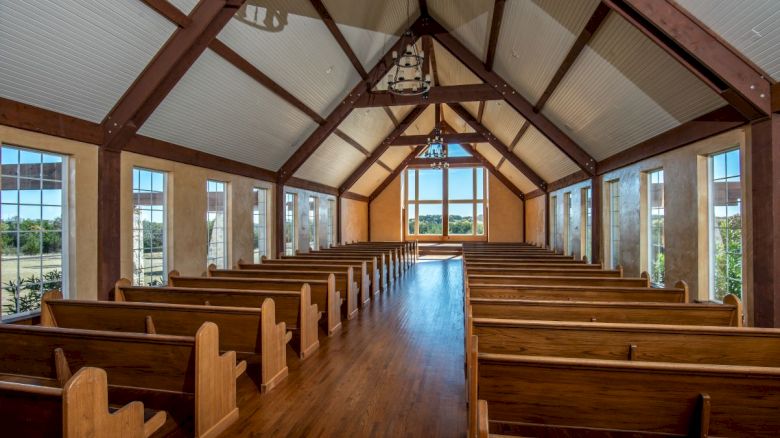 The image shows a wooden chapel interior with rows of pews, large windows, and a high ceiling, leading to a bright view outside.