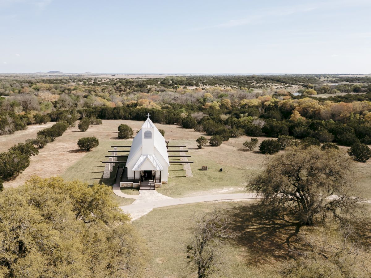 Aerial view of a small white chapel with a cross in a dry grassy landscape, surrounded by scattered trees and fields.