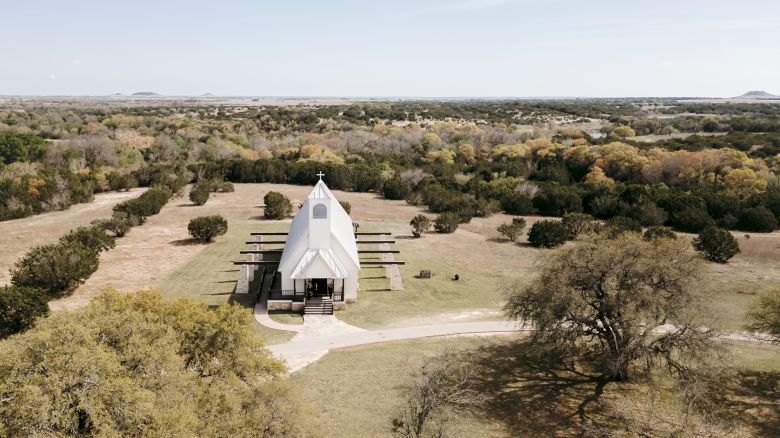 Aerial view of a small white chapel with a cross in a dry grassy landscape, surrounded by scattered trees and fields.