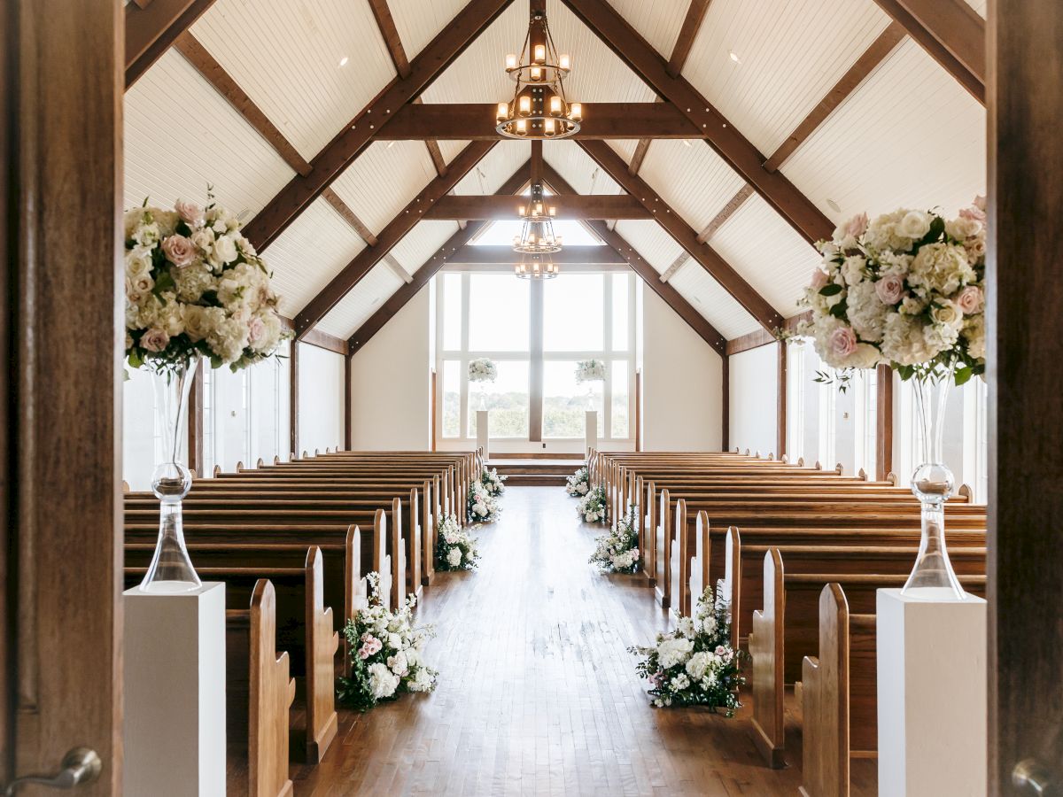 A decorated church aisle with wooden pews, floral arrangements, and hanging lantern-style chandeliers under a vaulted white ceiling.