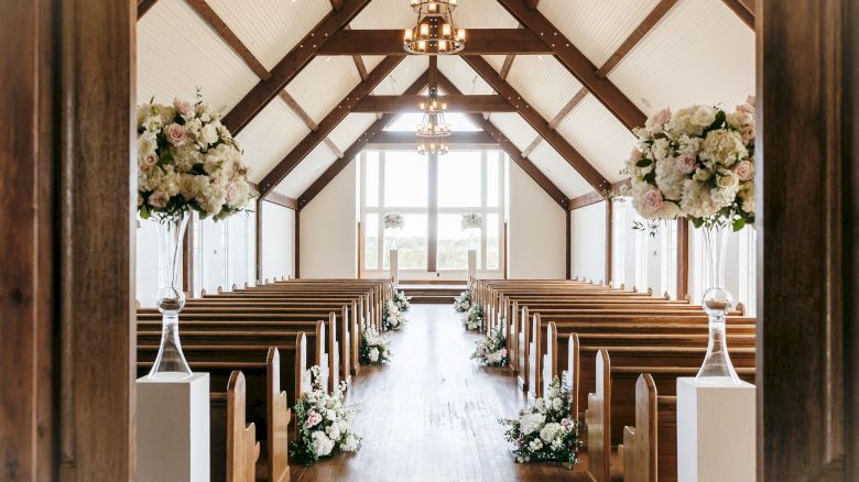 A decorated church aisle with wooden pews, floral arrangements, and hanging lantern-style chandeliers under a vaulted white ceiling.