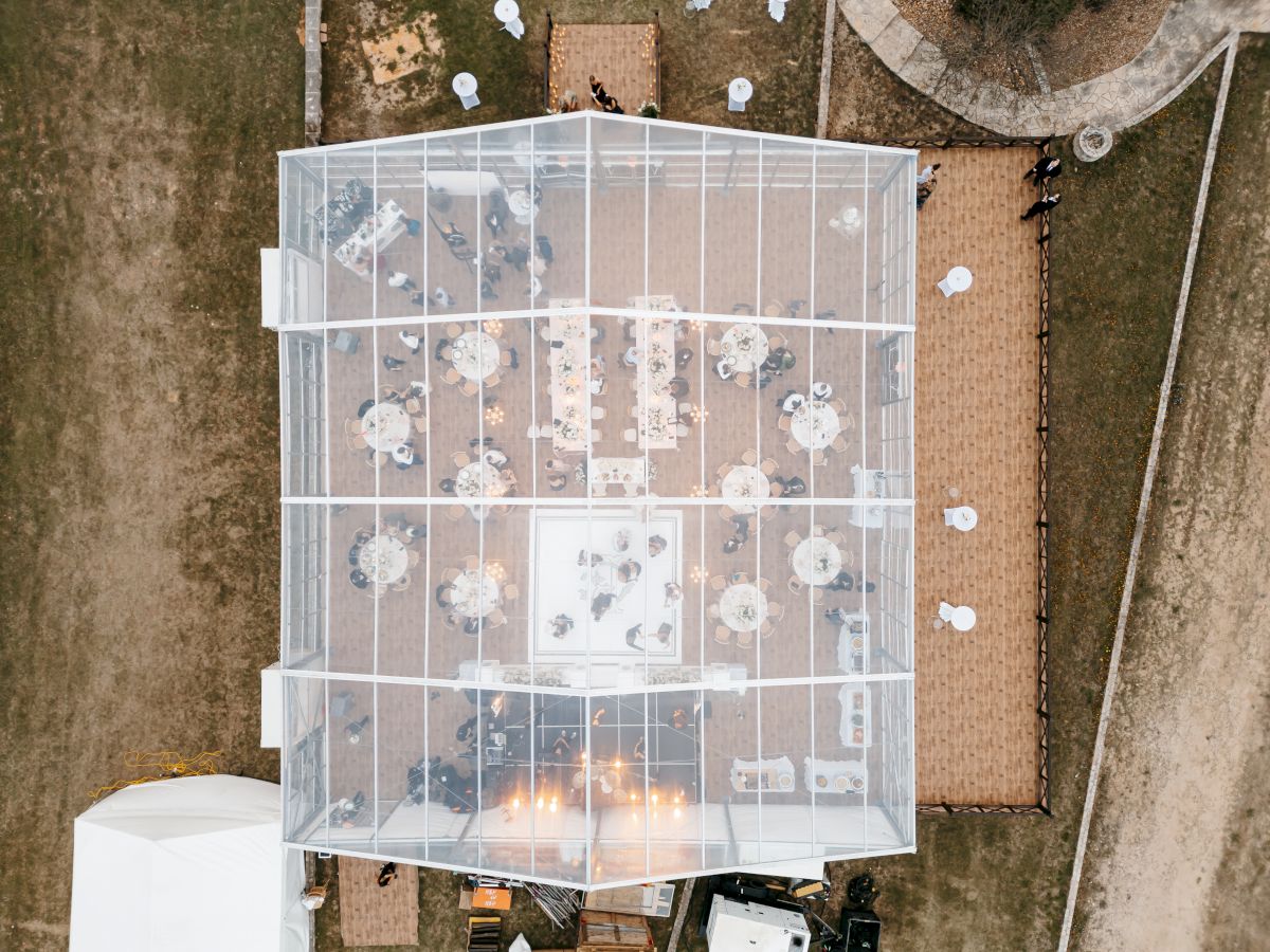 Aerial view of a wedding reception setup with a glass tent, round tables, candles, and guests seated inside on a lawn.