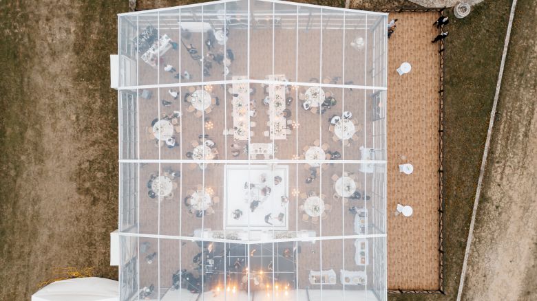 Aerial view of a wedding reception setup with a glass tent, round tables, candles, and guests seated inside on a lawn.